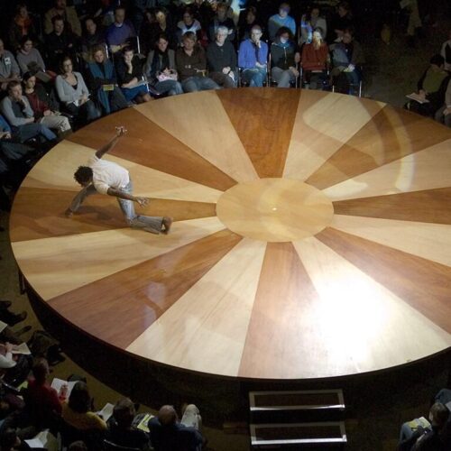 Ujęcie z góry na okrągłą drewnianą scenę. Na scenie tańczy czarnoskóry mężczyzna. Wkoło sceny zgromadzona jest liczna widownia.