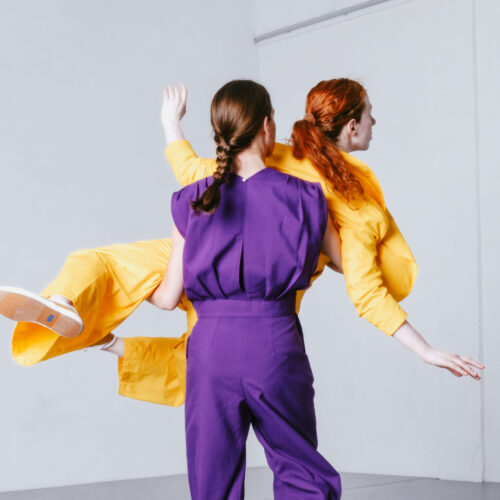 Kolorowa fotografia. Na jasnej sali prób dwie tańczące kobiety. Jedna z nich ubrana w fioletowy strój podnosi na rękach przed sobą drugą z nich ubraną na żółto. Fotografia jest dynamiczna.